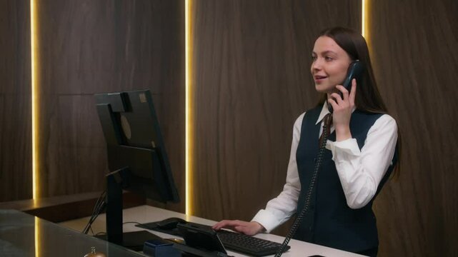 Female hotel receptionist in formal uniform answering phone call and typing on computer while working at front desk