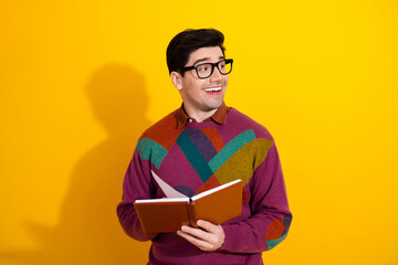 Cheerful man in a colorful sweater and glasses holding a notebook against a vibrant yellow...