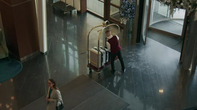 High angle shot of bellboy in uniform welcoming businesswoman entering hotel lobby, showing her way to checkin desk and assisting with luggage