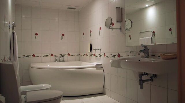 Sleek, Very Clean Hotel Bathroom with Large Corner Bathtub. Mirror, Toilet, Bidet, Sink, Tiles with Decorative Flowers, Towels, Soap, Glasses.
