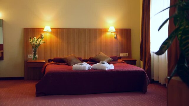 A Chic Hotel Bedroom Decorated in Red Tones. Huge Wooden Headboard. Lit Night Lights. Two White Robes and Slippers on the Bed for a SPA Visit.