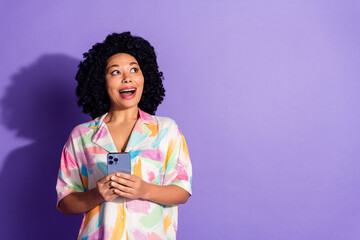 Young woman with curly hair holding smartphone, smiling against violet background, dressed in...