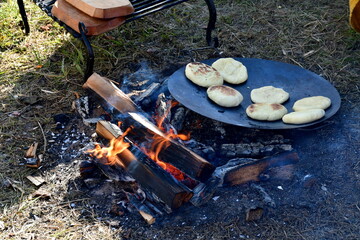 A close up on dumplings or shortbread cookies baked by using an iron pan laying in the firewood composed of logs, planks, and boards, seen next to an iron grate and a lush lawn in Poland