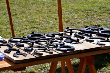 A close up on a set of horseshoes of various sizes and shapes made out of iron, brass, and steel, displayed on a wooden table or stand next to a lush lawn, meadow, or pastureland in Poland