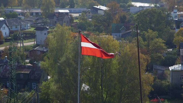 The Latvian Flag Flutters in the Wind High Above a Rural Village. The Flag of Latvia is Crimson Red with a Horizontal White Stripe in the Middle. It was Created in the 19th Century.