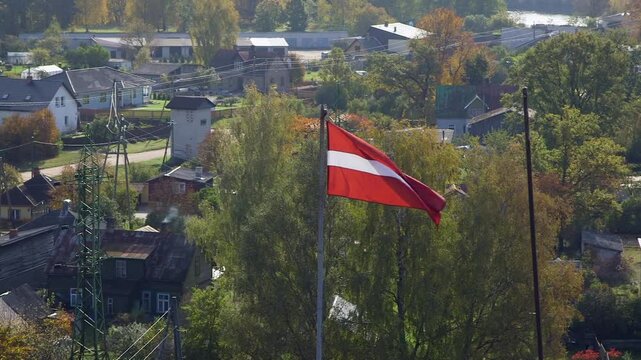 The Latvian Flag Flutters in the Wind High Above a Rural Village. The Flag of Latvia is Crimson Red with a Horizontal White Stripe in the Middle. It was Created in the 19th Century.