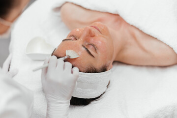 Cosmetologist cleaning face of a middle aged European woman with special gel before applying mask,...