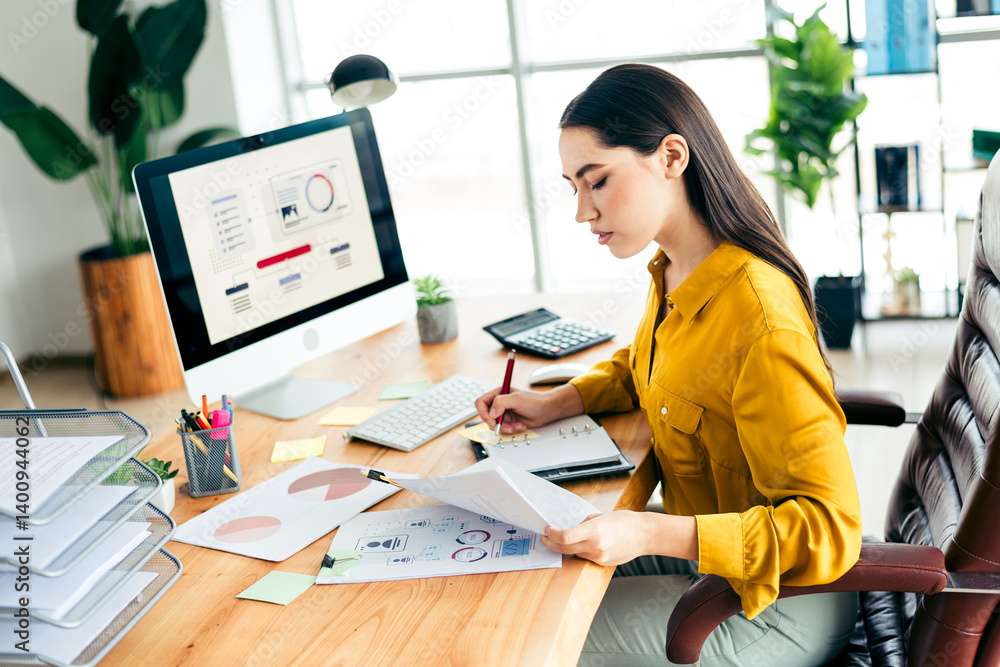 Poster skilled professional woman analyzing data at a modern office workspace with computer and documents d