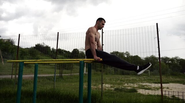 Strong man trains abdominal muscles raising legs on parallel bars at sports ground. Young shirtless sportsman exercising at nature. Sporty athlete working out at nature. Concept of sportive lifestyle