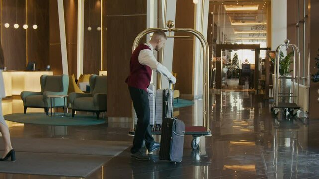 Bellhop in formal uniform greeting female guest near reception area in hotel lobby, giving her direction and helping with luggage