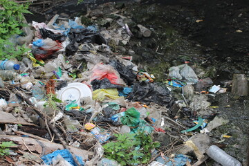 A powerful environmental image showing a polluted riverbank filled with plastic waste, bags,...