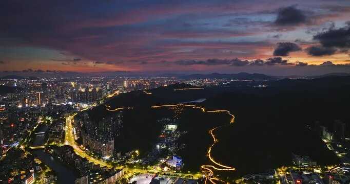 Aerial view of sprawling city adjacent to dark hills with a winding illuminated road under a dramatic colorful sunset sky in Zhuhai, China.
