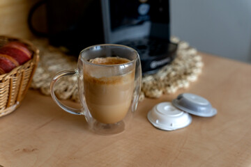 Glass of freshly brewed latte with foam on the background of capsule coffee maker in home kitchen.