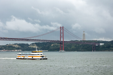 A scenic ferry crosses the Tagus River in Lisbon, Portugal, with the iconic 25 de Abril Bridge and Cristo Rei statue in the background under a dramatic cloudy sky.