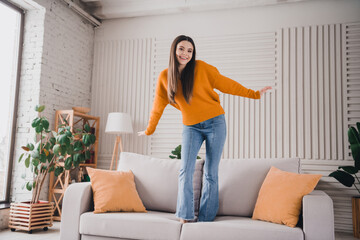 Young cheerful woman enjoying leisure time at home standing on a couch in a cozy living room with...