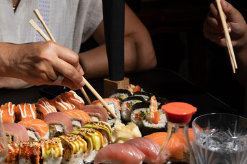 A table filled with various sushi pieces, including rolls and nigiri, with hands holding chopsticks, ready to eat. The scene captures a shared dining experience in a minimalist, elegant setting.