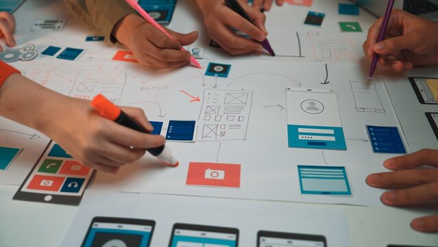 A group of creative individuals engaged in a productive design session for user interface layouts, utilizing colorful markers to annotate their sketches on a large paper. SACTR