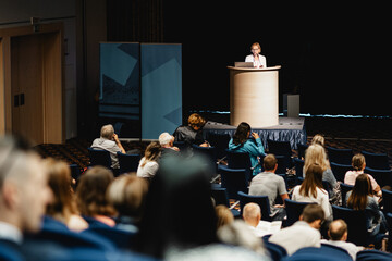 Female speaker giving a talk on corporate business conference. Unrecognizable people in audience at...