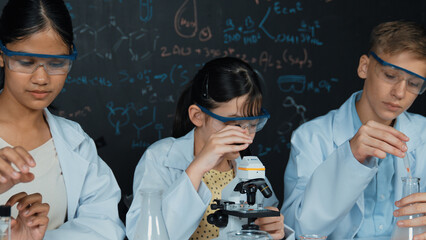 Smart teenager drop sample in microscope while girl inspect carefully at blackboard with chemistry...