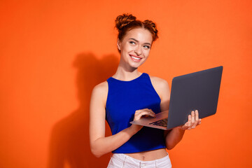 Young woman with a cheerful expression using a laptop against an orange background, wearing a...