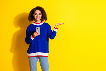 Young woman in casual colorful sweater holding smartphone and gesturing against a vibrant yellow...
