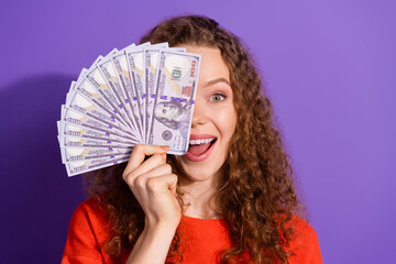 Smiling woman holding fan of dollar bills against purple backdrop, promoting finance and happiness