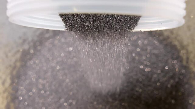 Dark abrasive diamond powder falling from plastic container in controlled flow, Macro view of precise raw material handling in industrial tool production environment