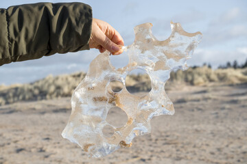 
A hand holding an irregularly shaped piece of ice with holes against a sandy landscape background.