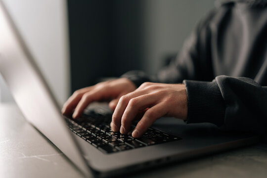 Close-up hands of unrecognizable programmer male typing on laptop keyboard, immersed in software development or coding, sitting at table by window in dark room. Concept of remote job, freelance.