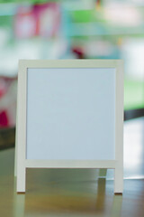 Blank white frame board standing on a wooden surface with a colorful blurry background. Ideal for mockups, text design, or marketing visuals.