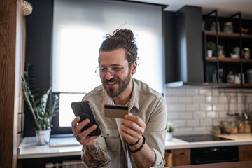 Freelancer smiling and buying online with credit card and smartphone in his modern kitchen