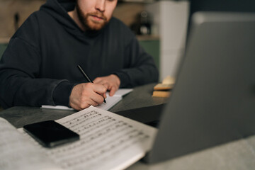 Cropped shot of music composer writing melodic notes in home studio, sitting at table with laptop, smartphone and sheet music scattered across workspace, merging artistic expression with digital tools