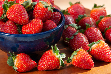 Fresh strawberries in a bowl with scattered berries on a wooden table during sunny daylight