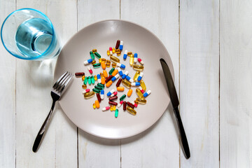 Mixed pills on plate with fork and knife. Various colorful capsules and tablets are scattered on a plate, styled like a meal, with cutlery and a glass of water.