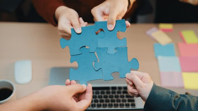 Two hands connect vibrant puzzle pieces on a table with a laptop and colorful sticky notes, symbolizing teamwork and strategy in a collaborative office setting. SACTR