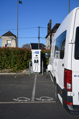 An electric van is charging at a charge stationin a hedged car park. A pictogram authorizes electric vehicles. Rear view. Vertical picture.