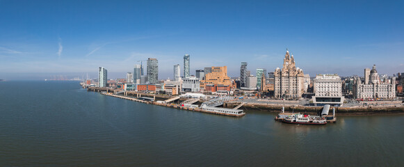 Naklejka premium Aerial view of Liverpool's waterfront featuring the Three Graces, modern skyscrapers, the River Mersey, and a ferry docked near the shore.