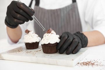 Confectioner decorating tasty cupcake with chocolate shaving at white wooden table in kitchen, closeup
