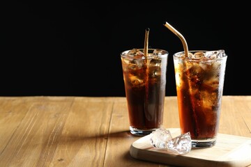 Refreshing cola with ice cubes and straws in glasses on wooden table against black background. Space for text