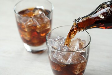 Pouring refreshing cola from bottle into glass at light grey table, closeup