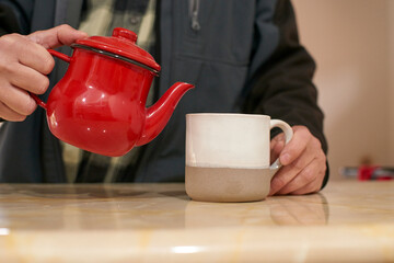 Man pouring steaming tea from vintage red teapot into ceramic mug, savoring peaceful morning moment at kitchen counter
