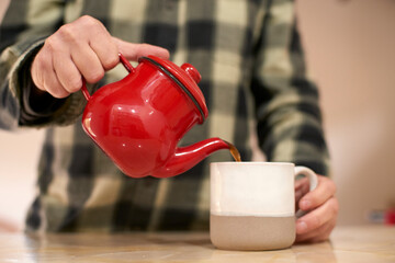 Man in plaid shirt pouring steaming tea from vintage red teapot into two-tone ceramic mug, savoring peaceful morning moment at home