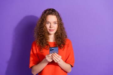 Young woman with curly hair holding a smartphone in front of a violet background, wearing an orange...