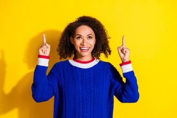 Cheerful Young Woman Pointing Upward Against a Vibrant Yellow Background in a Stylish Blue Sweater