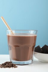 Tasty chocolate milk with shavings and pieces on white table against light blue background, closeup