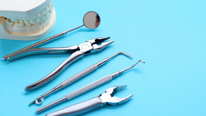 Professional metallic dentist tools including mirror, forceps, scaler probe and jaw model arranged on a clean light blue background under bright studio lighting, high angle view.