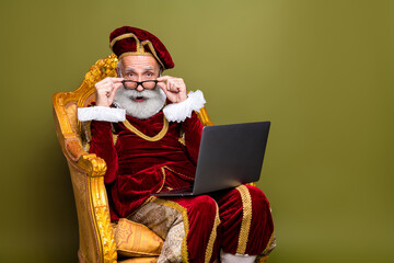 Elderly nobleman in medieval costume and beret sitting on royal chair with laptop, against khaki...