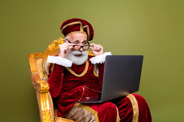 Elderly man in royal renaissance attire using a modern laptop on a golden chair against khaki...