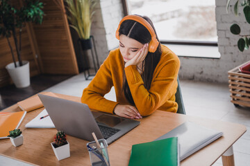 Young girl wearing orange headphones, studying at home with her laptop on a wooden desk in a modern room with natural daylight