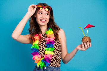 Young woman wearing a colorful lei and sunglasses holding a coconut drink while smiling against a blue background. © deagreez